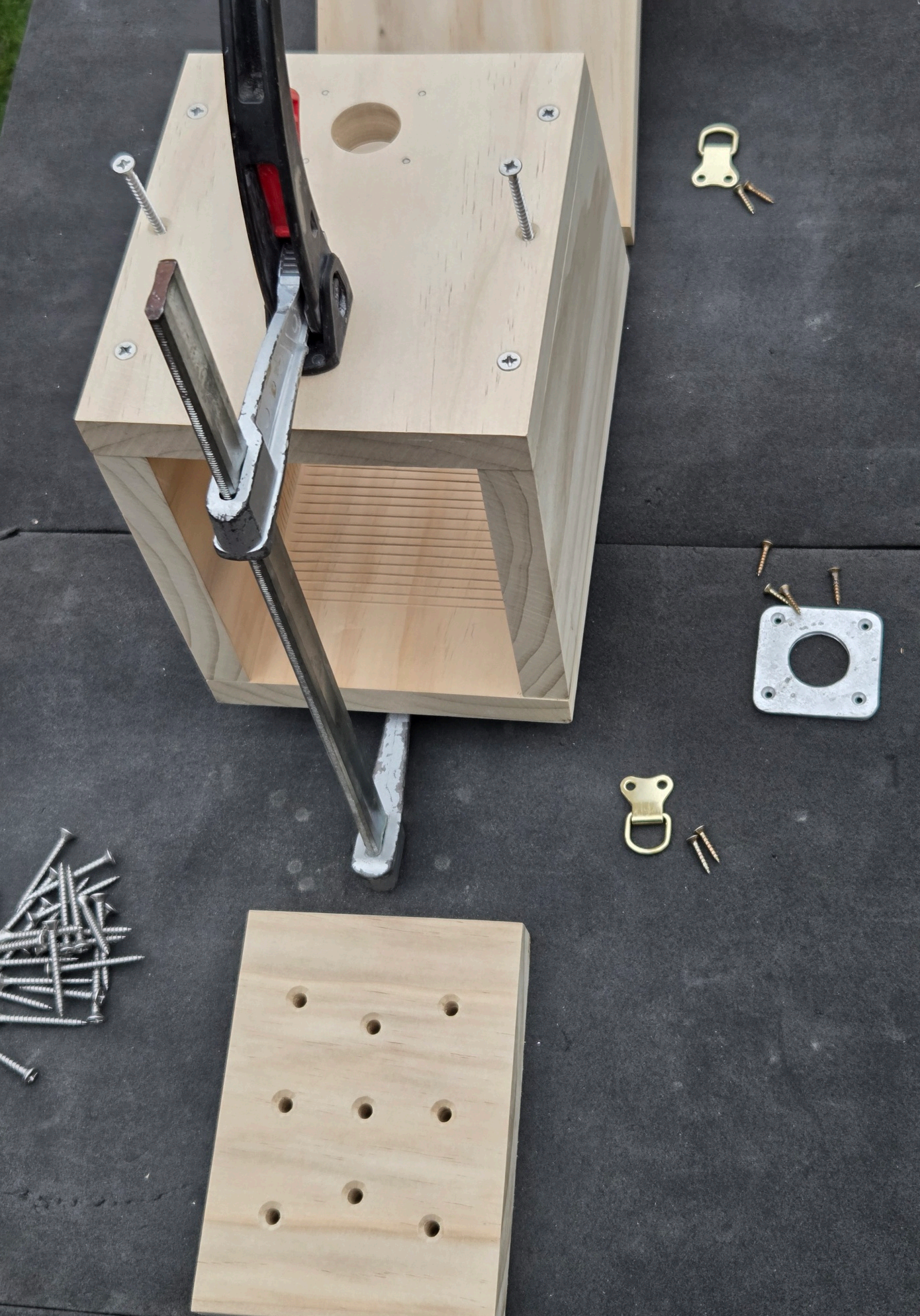 Wooden box frame being held together with a clamp, screws and hardware laid out nearby, floor piece with drainage holes below